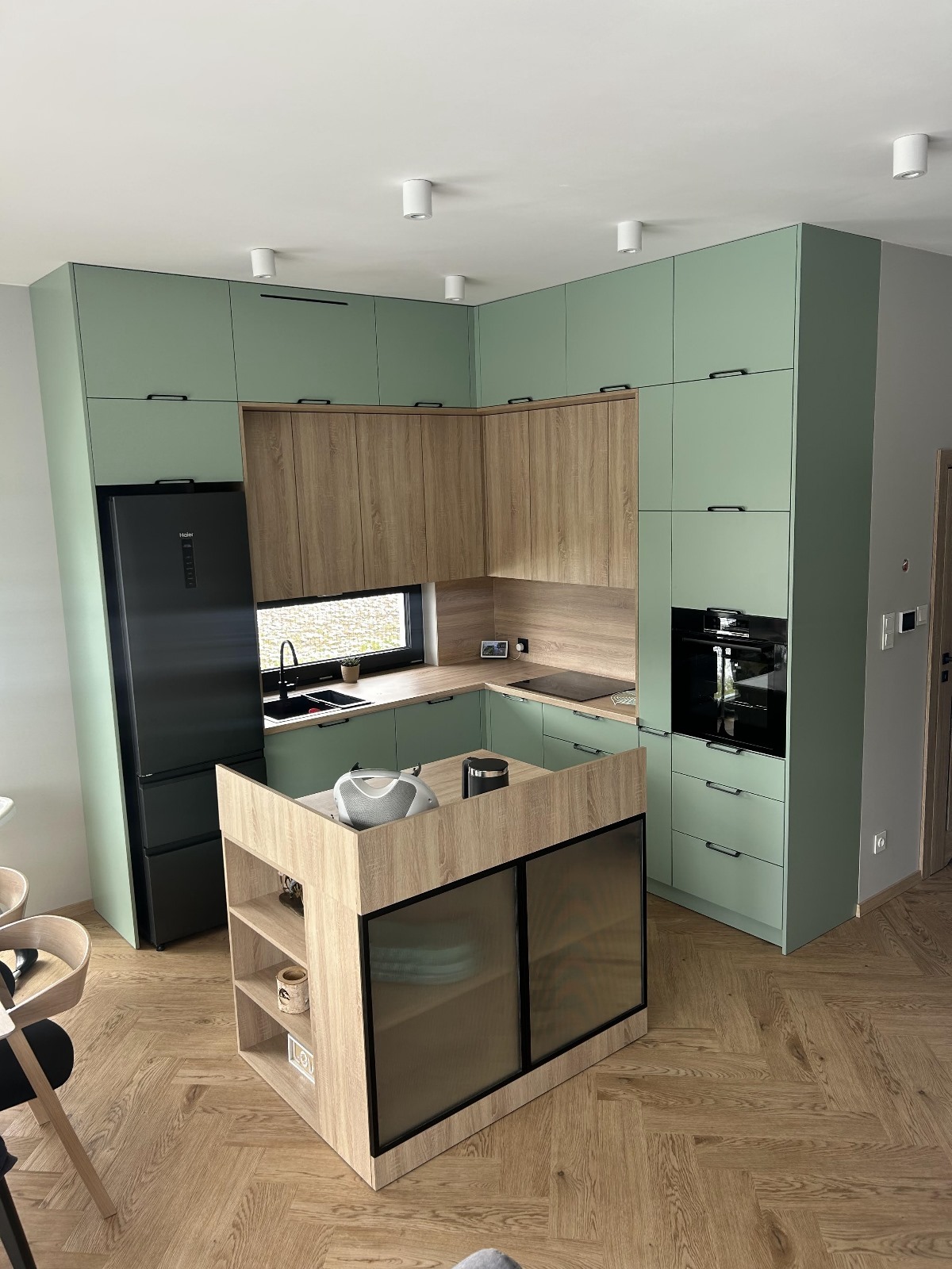 Nowoczesna kuchnia w odcieniach zieleni i drewna z wyspą, czarnym sprzętem AGD i parkietem ułożonym w jodełkę. Widok na całe pomieszczenie, oświetlenie punktowe.