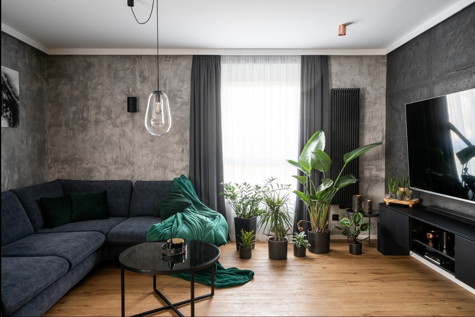 Stylowy salon z drewnianą podłogą, szarą sofą narożną, czarnym stolikiem i licznymi roślinami doniczkowymi. Ściana z efektem betonu i czarny grzejnik.