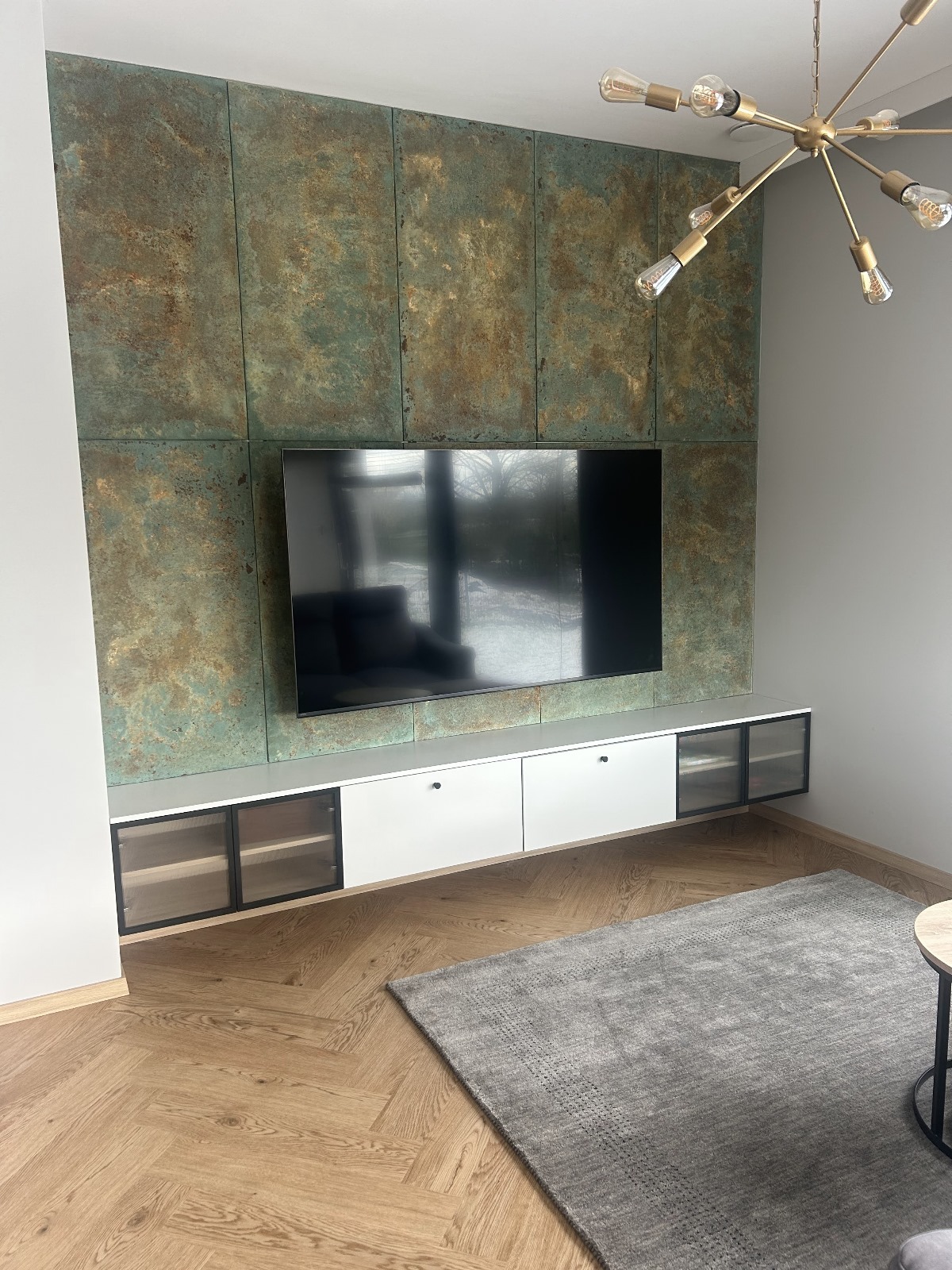 Salon z parkietem w jodełkę, ścianą z dekoracyjnych paneli i szafką RTV pod telewizorem. Całość oświetlona nowoczesną lampą.