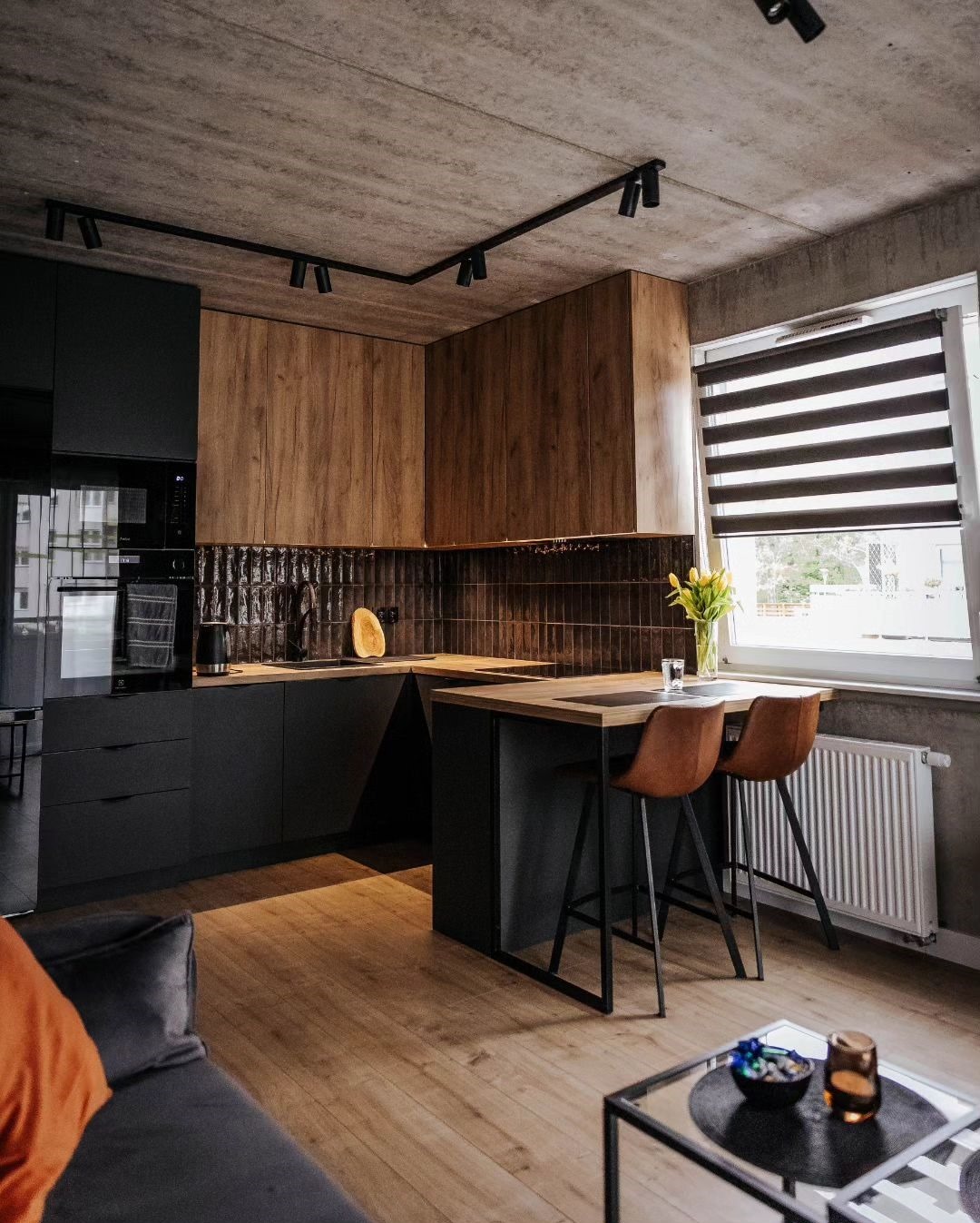 Nowoczesna kuchnia z drewnianymi frontami, czarnymi szafkami i wyspą. Parkiet w jasnym odcieniu. Oświetlenie szynowe na betonowym suficie. Minimalistyczny design.