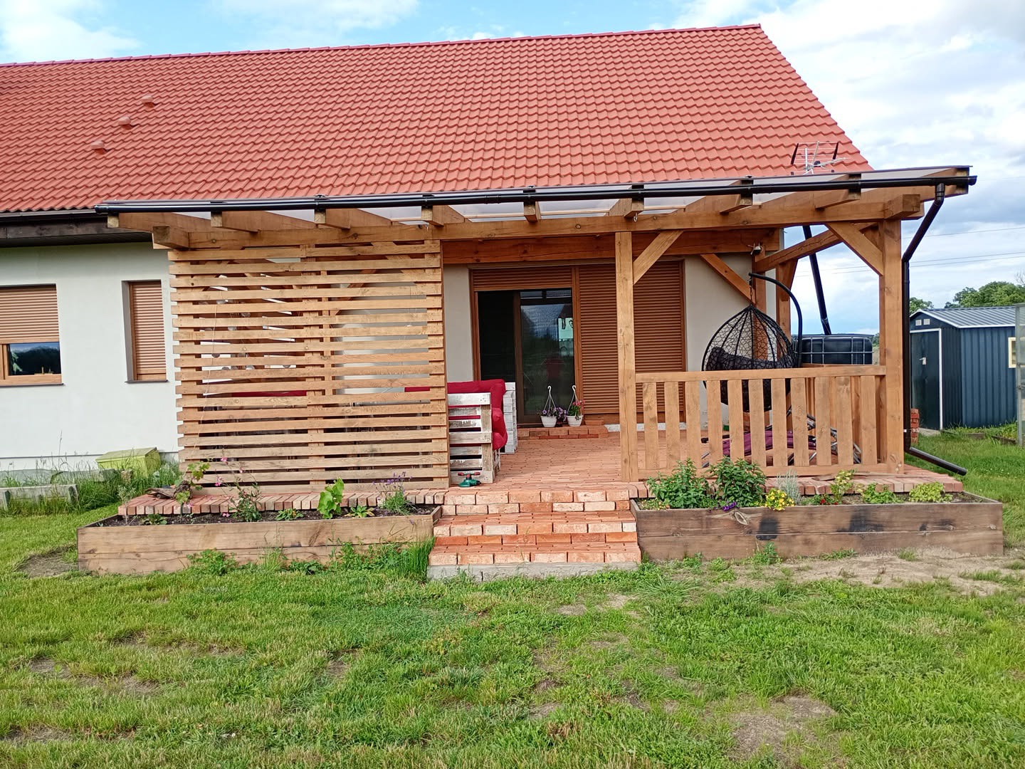 Drewniana pergola z zadaszeniem przy domu z czerwoną dachówką. Taras wyłożony cegłą, z drewnianymi skrzyniami na rośliny. Widoczne krzesło wiszące.