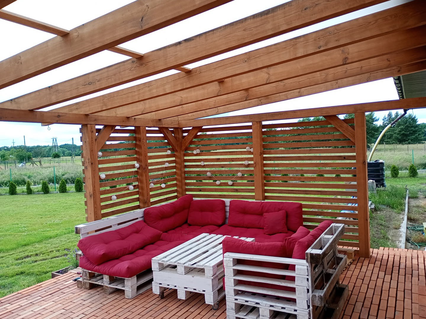 Zadaszona, drewniana konstrukcja z ażurowymi ścianami, oświetlona sznurem lampek. Wewnątrz komplet wypoczynkowy z palet i czerwonymi poduchami na ceglanym tarasie.
