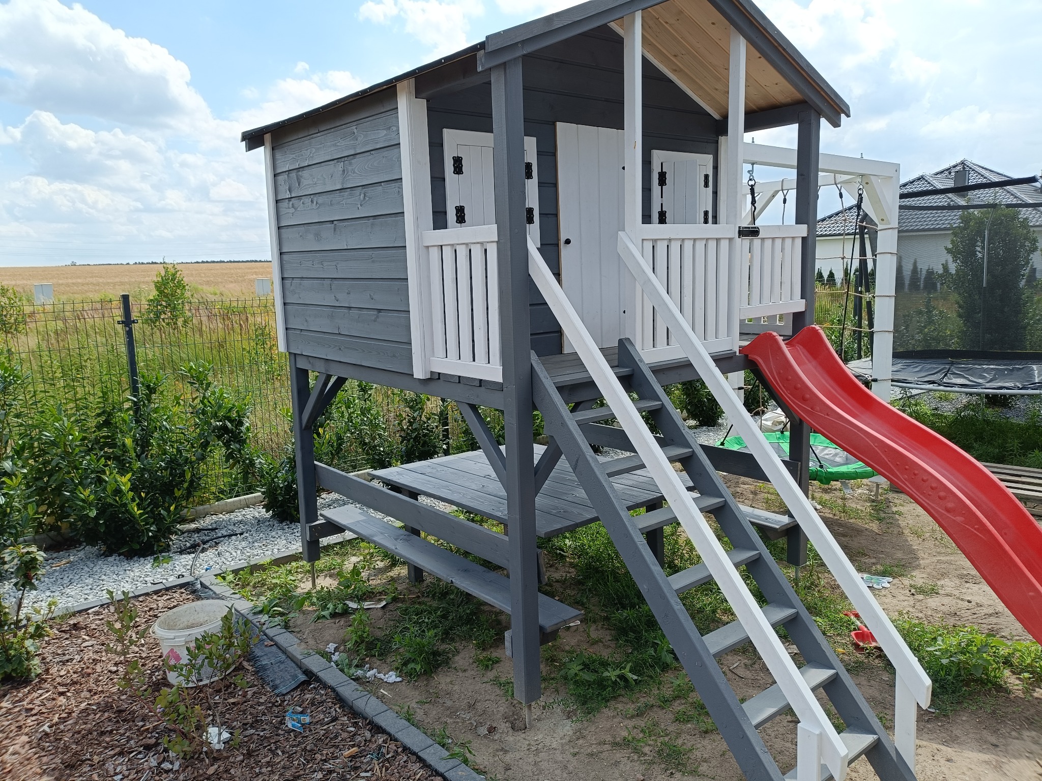 Szara, drewniana konstrukcja placu zabaw z białymi akcentami, schodami i czerwoną zjeżdżalnią, ustawiona w ogrodzie na tle zieleni i pola.