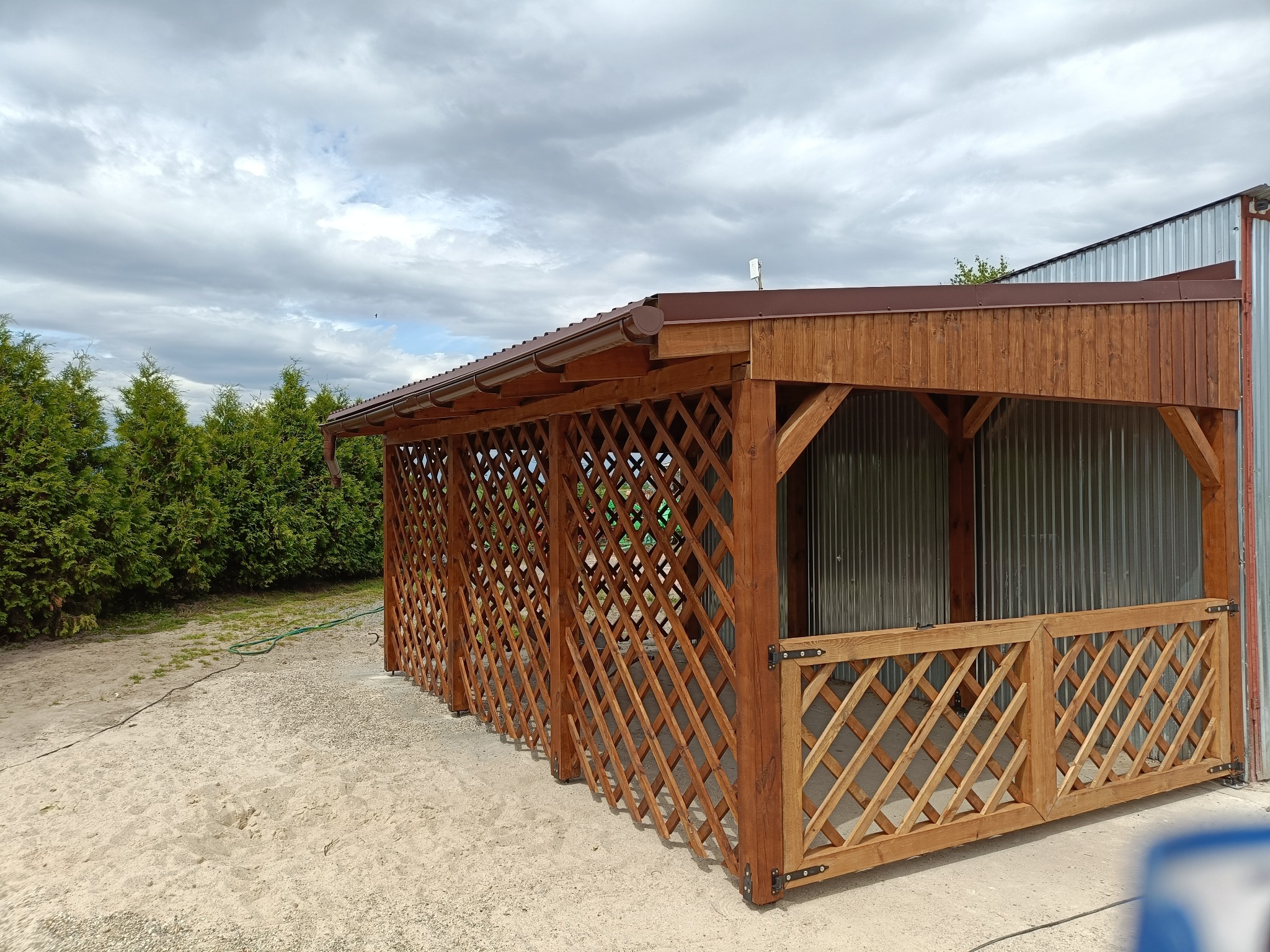 Drewniana altana z kratownicą i brązowym dachem, widoczna z zewnątrz, na tle zieleni i pochmurnego nieba. Konstrukcja częściowo osłonięta blachą falistą.