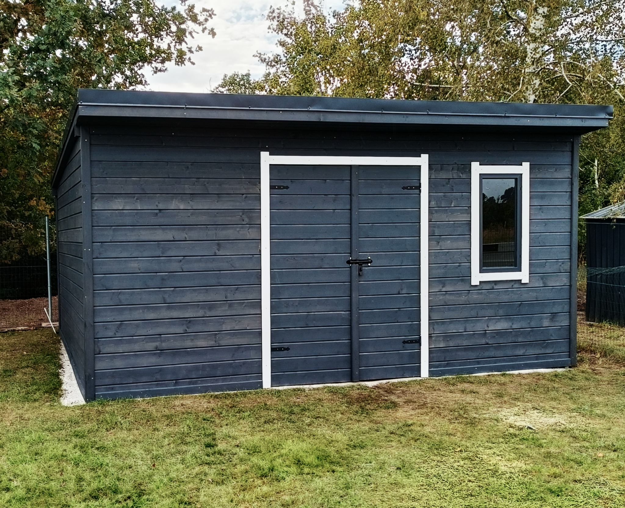 Nowoczesna, grafitowa altana ogrodowa z drewnianą konstrukcją i białymi obramowaniami drzwi i okna, na tle zielonego trawnika i drzew.