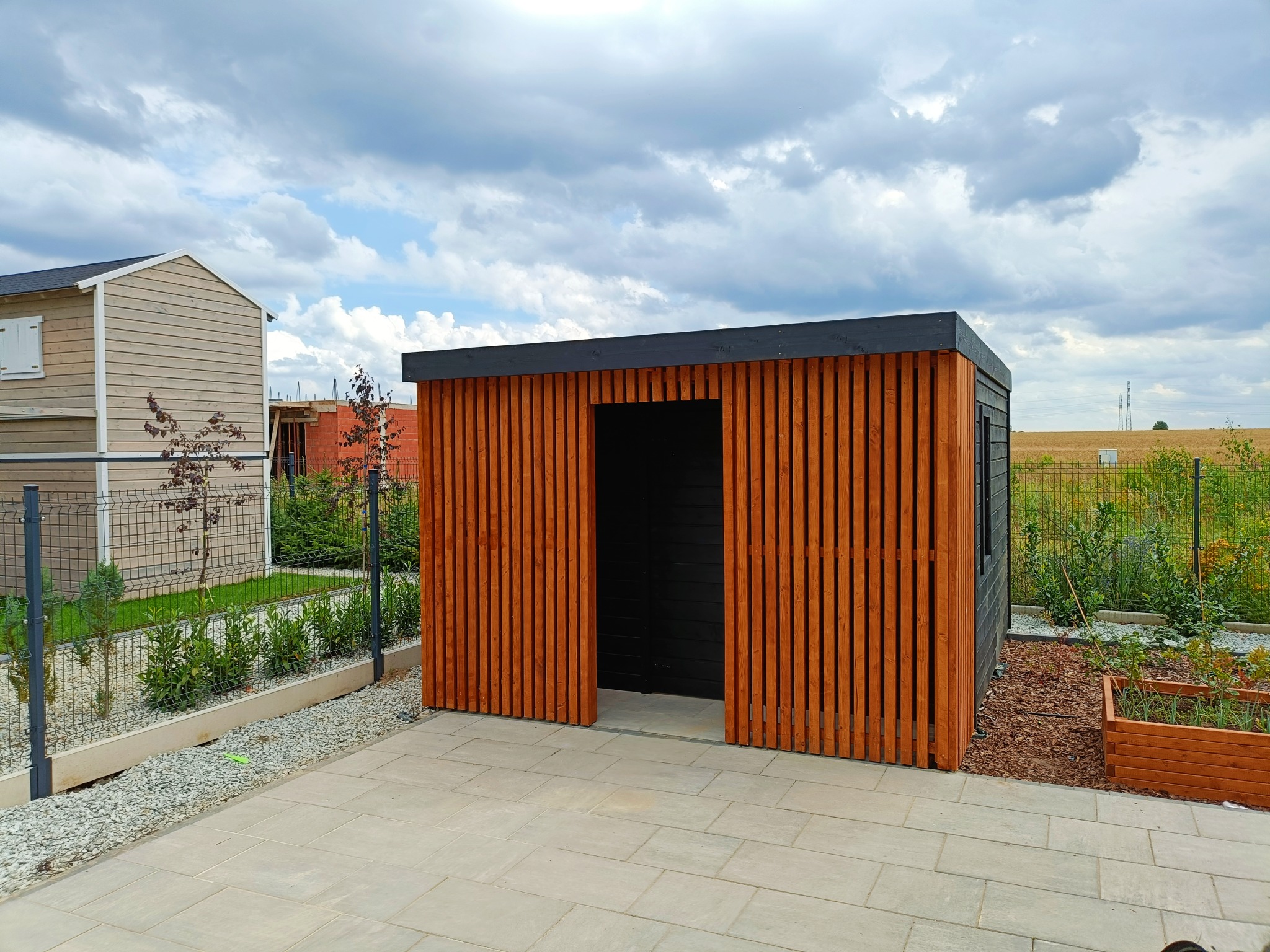 Nowoczesna, drewniana altana z otwartym wejściem, pomalowana na brązowo-czarno, na tle kostki brukowej i zieleni. Widok na sąsiedni budynek i pole.