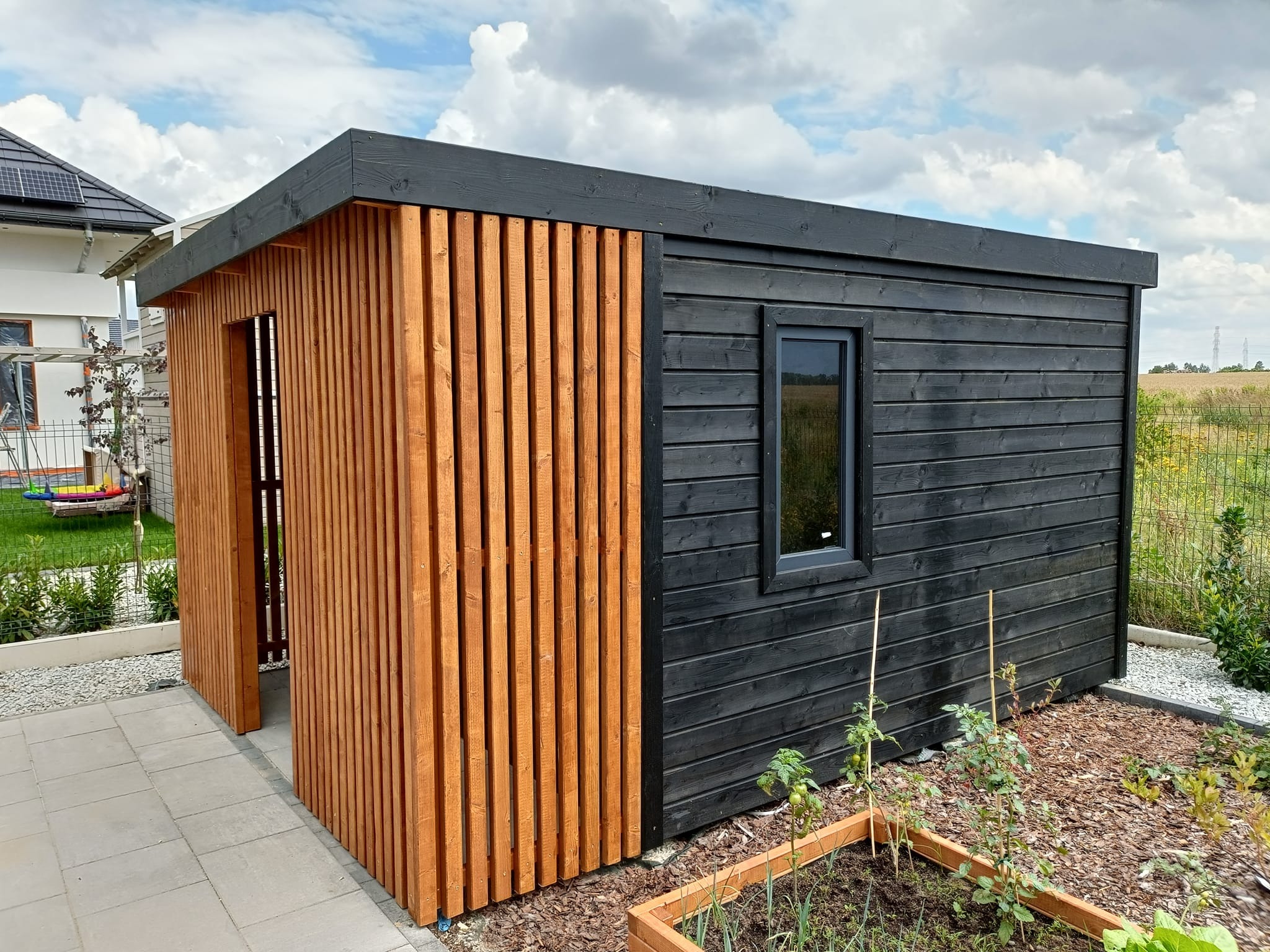 Nowoczesna, czarno-brązowa altana ogrodowa z drewna, z oknem i ażurową ścianą, na tle ogrodu i błękitnego nieba z chmurami.