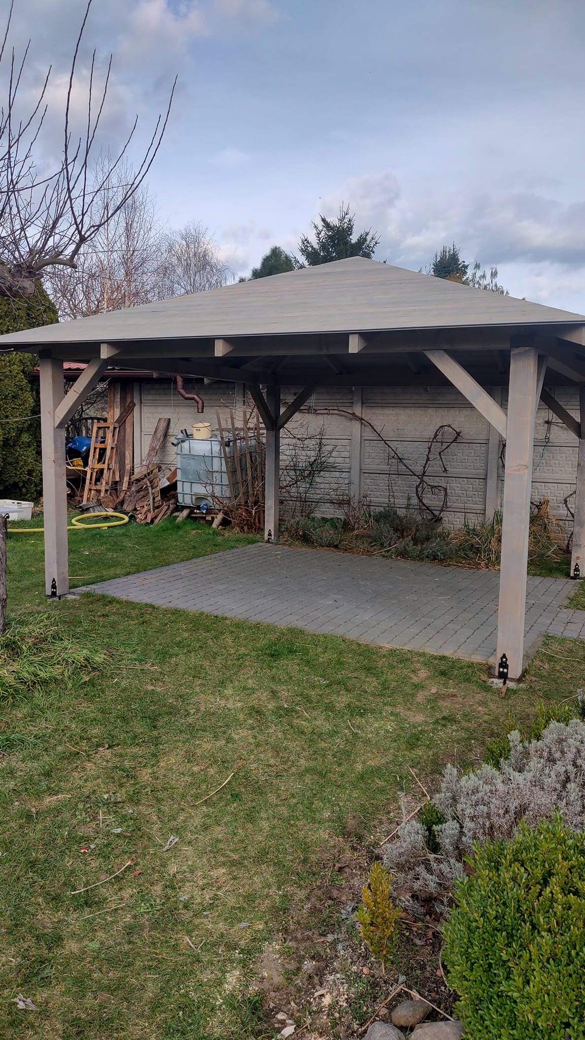 Drewniana pergola z szarym dachem na tle betonowego ogrodzenia i zielonego trawnika. Widoczne elementy konstrukcyjne i kostka brukowa pod pergolą.