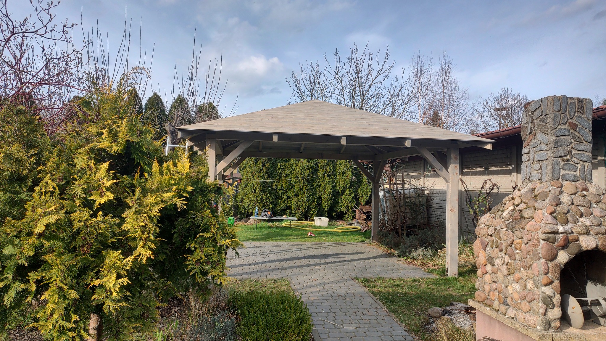 Drewniana altana z szarym dachem w ogrodzie w Prochowicach. Widoczna kamienna ściana budynku i ścieżka z kostki. Zieleń drzew i krzewów w tle.