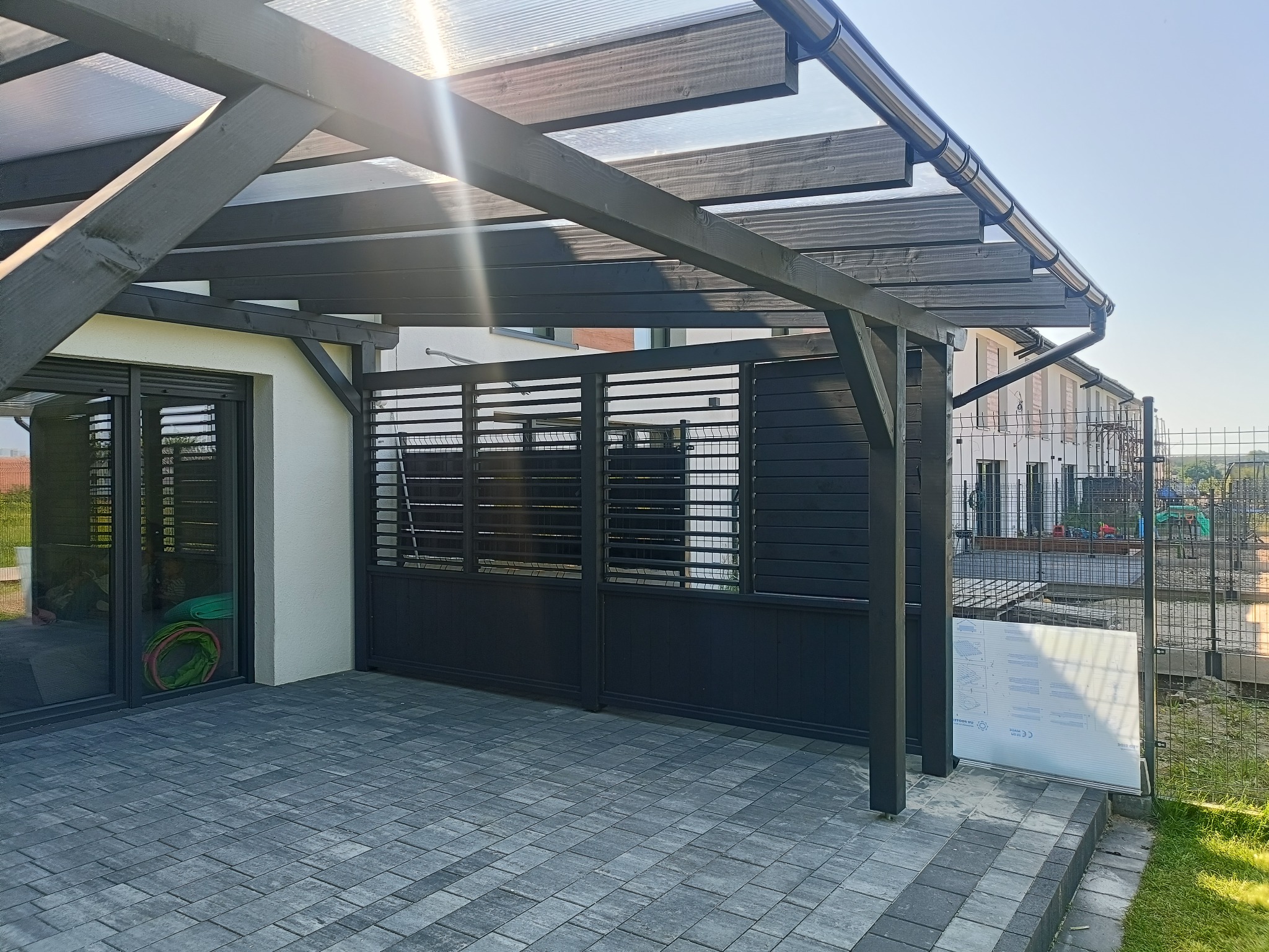 Nowoczesna, grafitowa pergola z zadaszeniem z tworzywa sztucznego, przylegająca do budynku. Widoczna ażurowa ścianka i kostka brukowa na podłożu.
