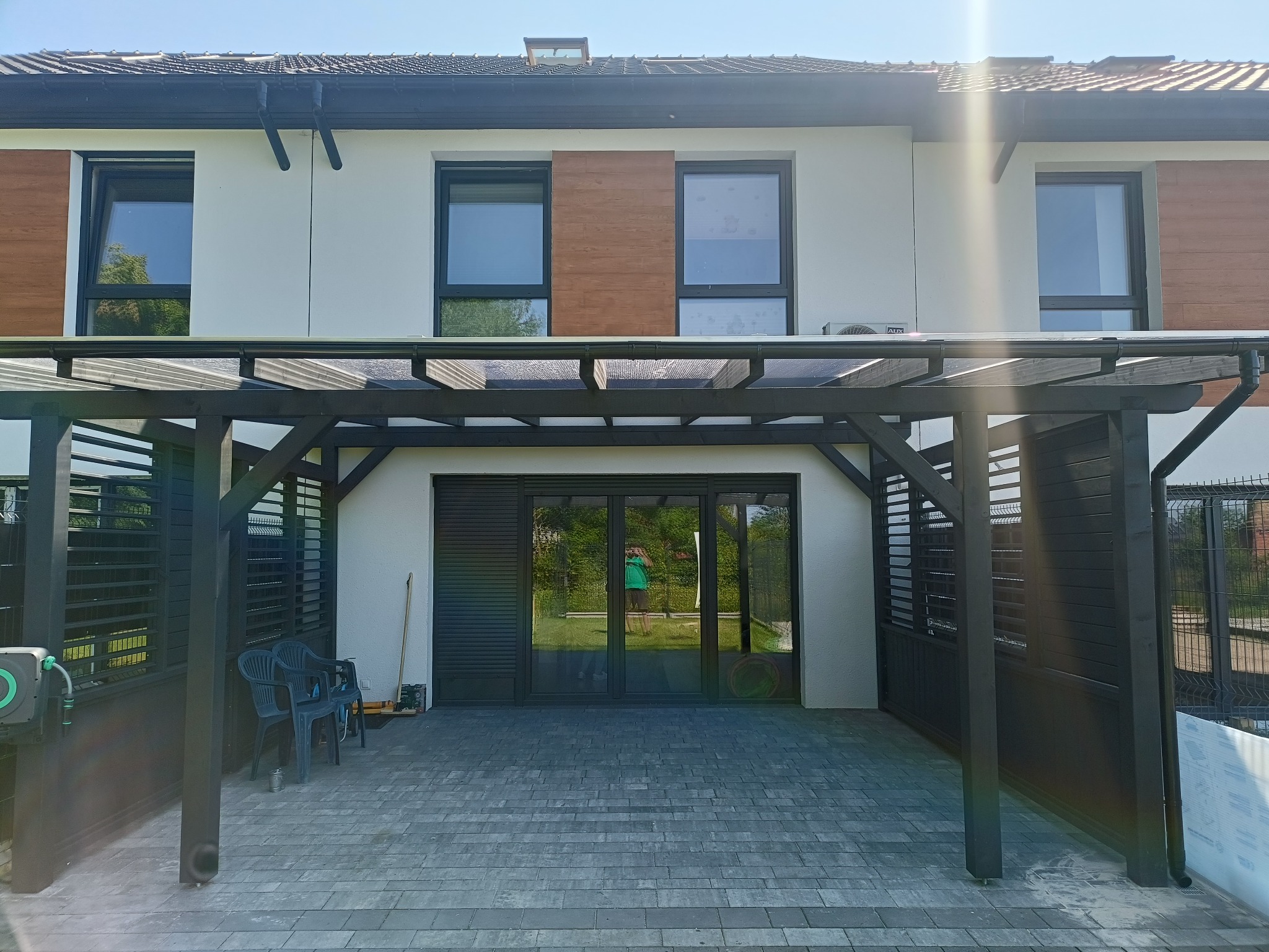 Nowoczesna pergola z ciemnego drewna przylegająca do domu, z widocznym zadaszeniem, szarym brukiem i elementami ogrodzenia. W tle budynek z jasną elewacją i ciemnymi oknami.