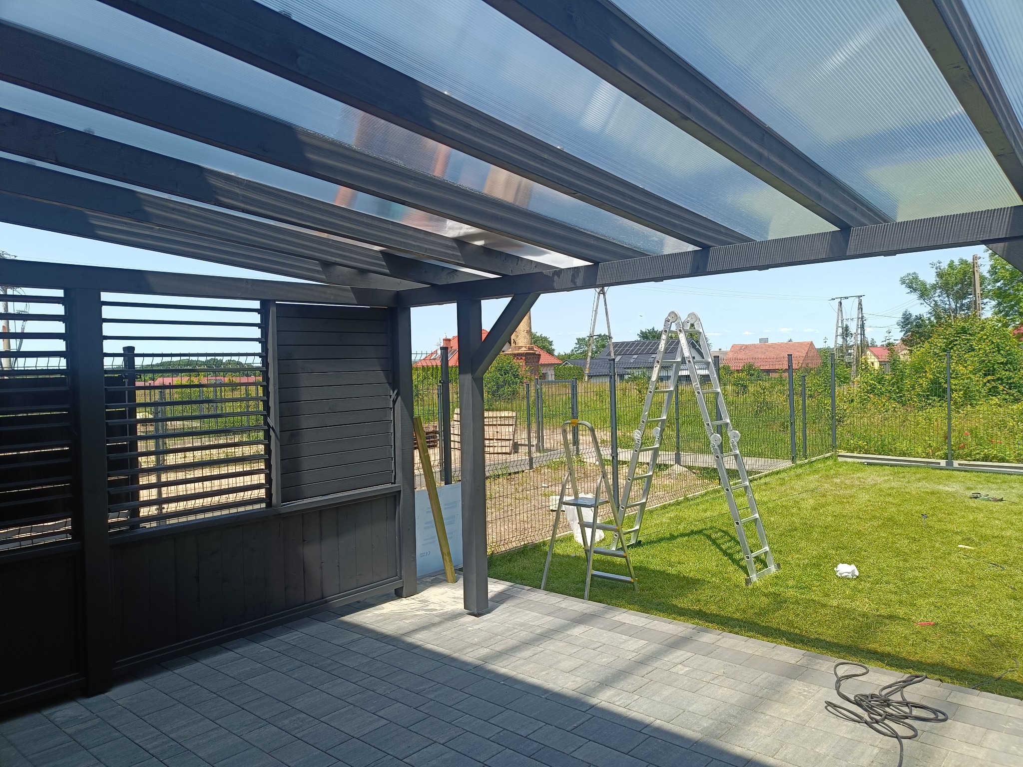 Zadaszenie tarasu z szarego drewna i przezroczystego tworzywa, widok z zewnątrz, drabina oparta o konstrukcję, trawnik, ogrodzenie z siatki.