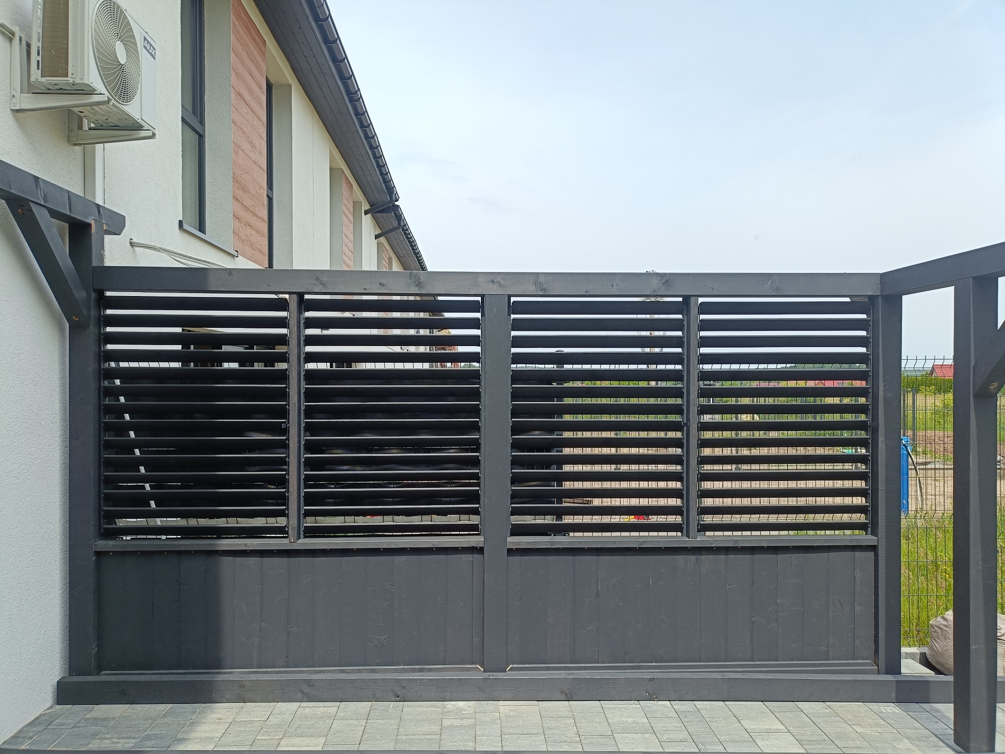 Nowoczesna, szara pergola z lamelami, przylegająca do białego budynku z klimatyzacją. Widoczne szare płytki tarasowe i zielona roślinność w tle.