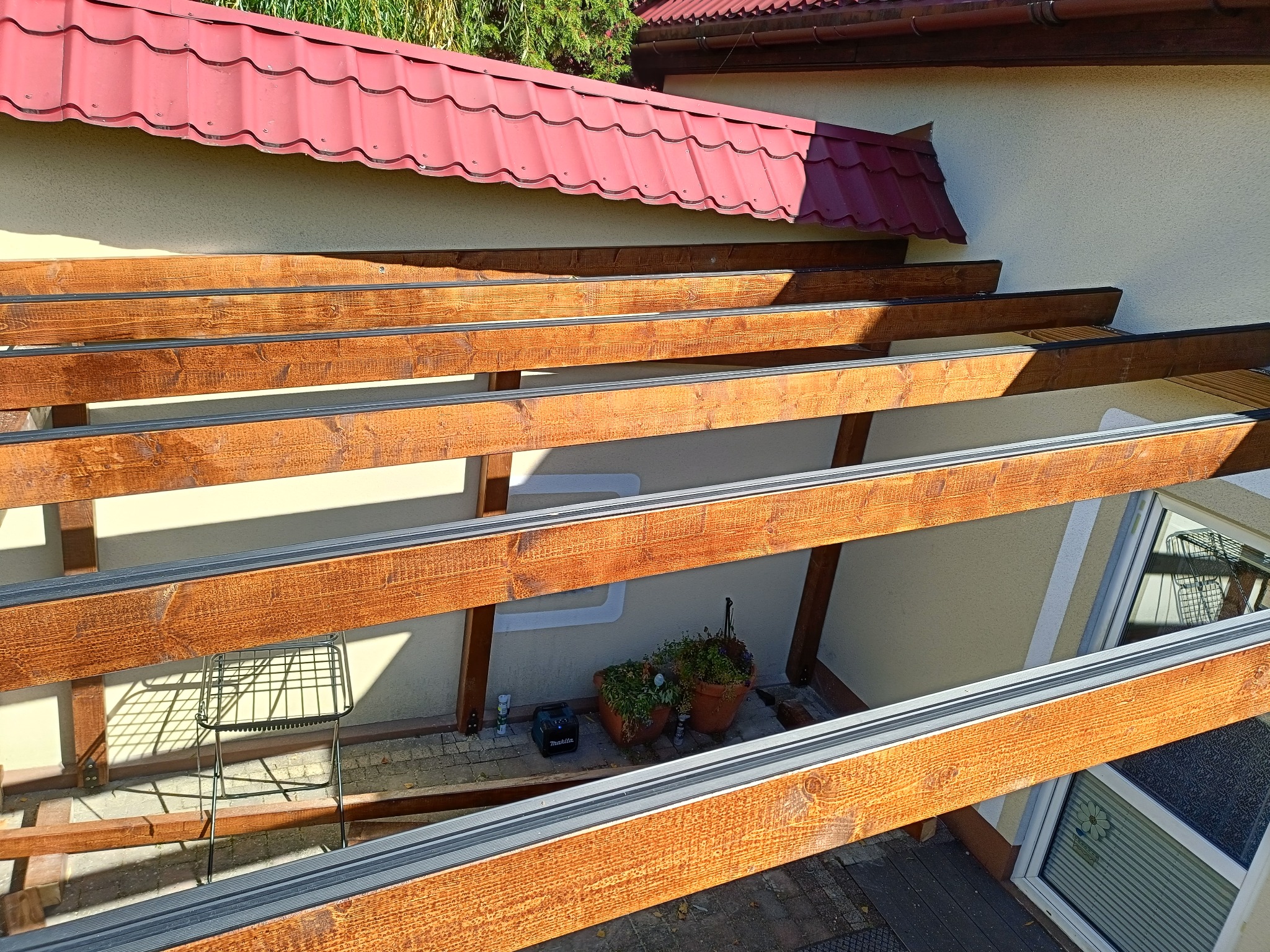 Drewniana konstrukcja z brązowych belek i szarych profili, prawdopodobnie w trakcie budowy z widocznym fragmentem dachu i ścian budynku w tle.