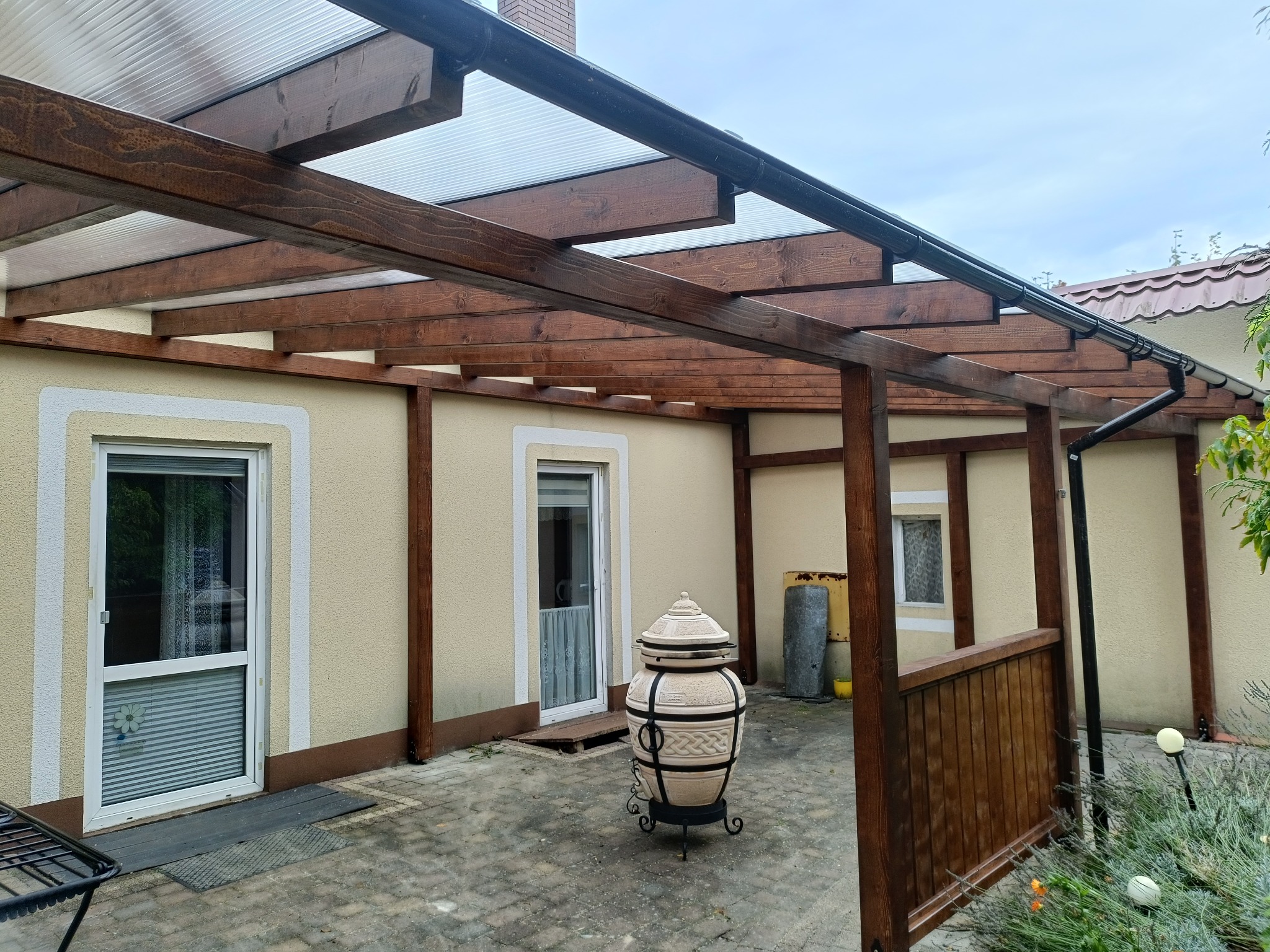 Zadaszona, drewniana pergola przy budynku z dwoma oknami i drzwiami. Na pierwszym planie dekoracyjny piec tandoor na brukowanym patio.