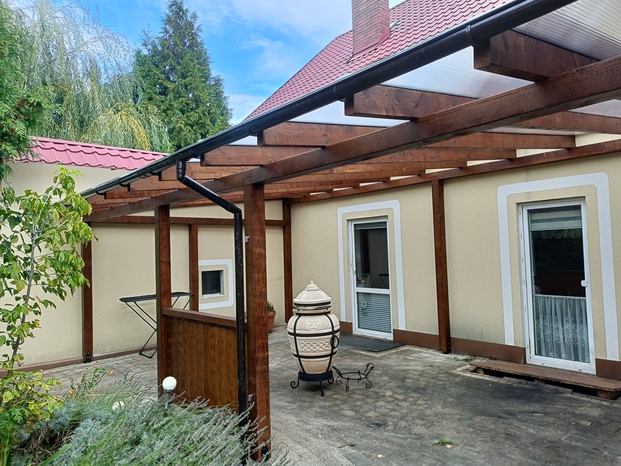 Drewniana konstrukcja z przezroczystym zadaszeniem przylegająca do budynku. W tle widoczny ogród i fragment dachu. Na pierwszym planie piec Tandoor.