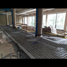 KM Instal Michał Kozyra - Rozłożona instalacja ogrzewania podłogowego z rurami na izolacji, widoczne okna i filary. Budynek w stanie surowym.
