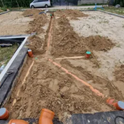 Instalacja kanalizacyjna w gruncie na działce budowlanej, widoczne pomarańczowe rury i studzienki. W tle dom jednorodzinny i zaparkowany van.