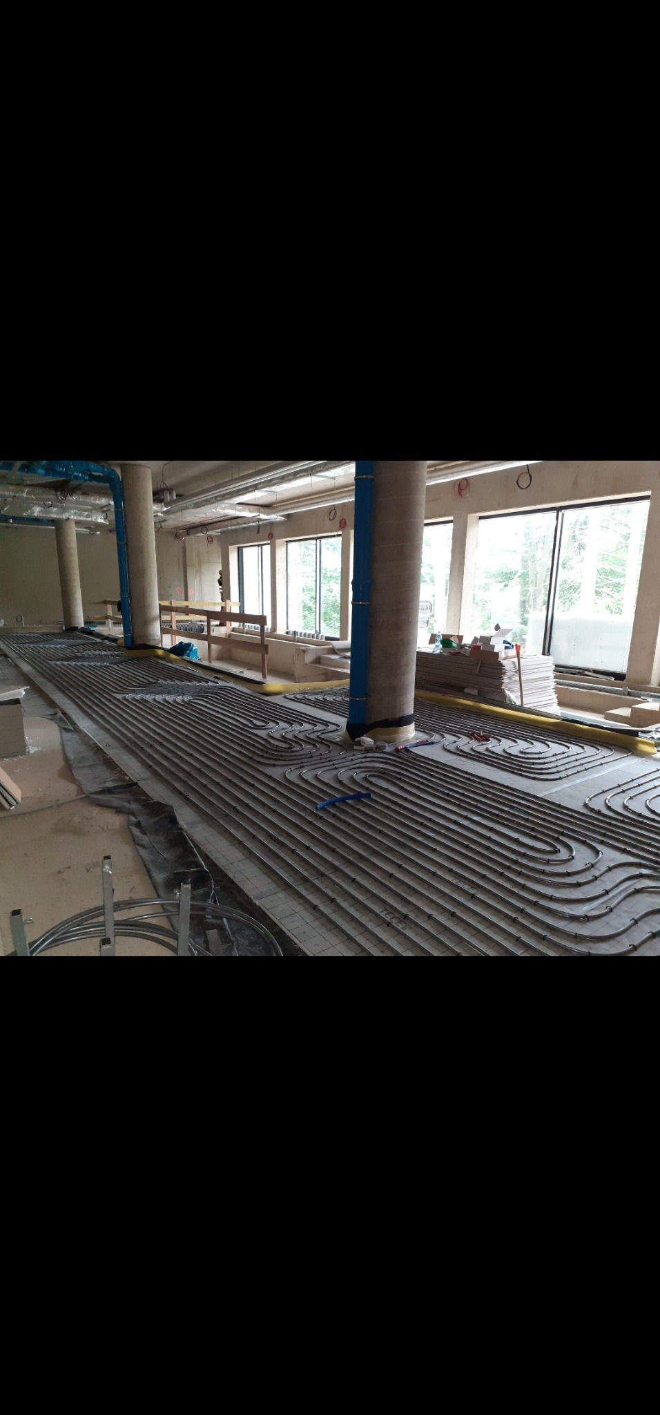 Rozłożona instalacja ogrzewania podłogowego z rurami na izolacji, widoczne okna i filary. Budynek w stanie surowym.