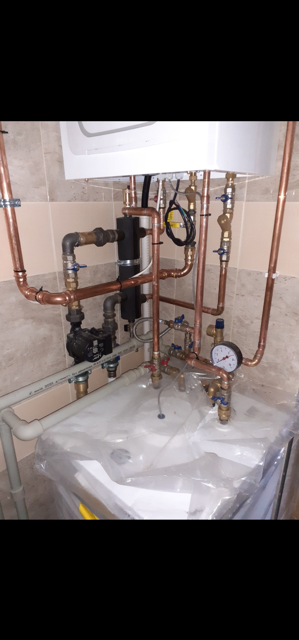 Nowa instalacja pieca gazowego z miedzianymi rurami, zaworami i manometrem, osłonięta folią ochronną. Widoczne elementy pompy obiegowej i izolacji termicznej.