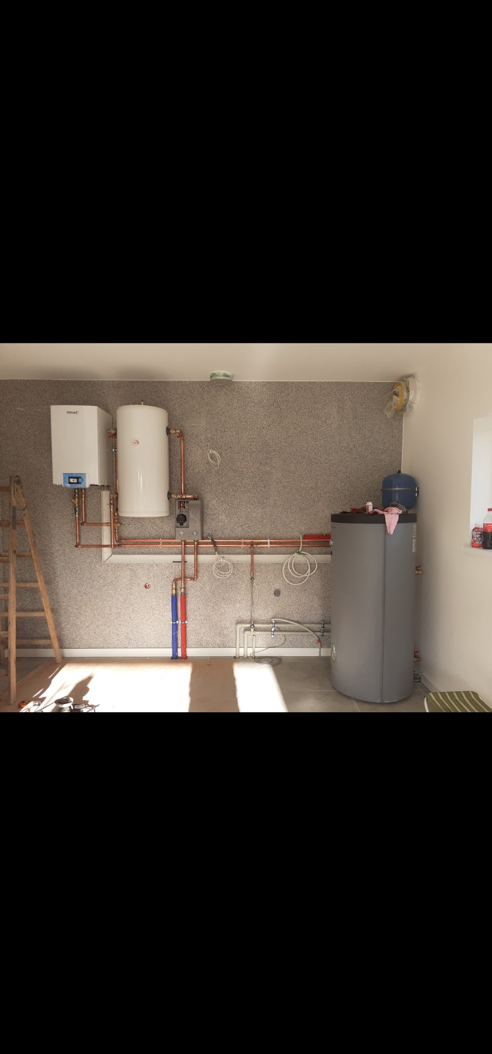 Nowoczesna kotłownia z białym piecem gazowym, zbiornikiem i miedzianą instalacją na tle ściany o fakturze baranka. Widoczna drabina i akcesoria na zbiorniku.