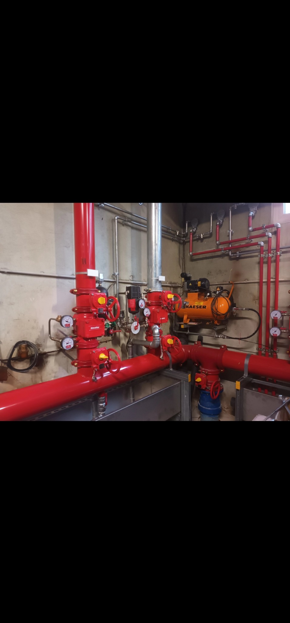 Czerwona instalacja rur z zaworami i manometrami w pomieszczeniu technicznym. Widoczny kompresor marki Kaeser. Rury prowadzone wzdłuż ściany.