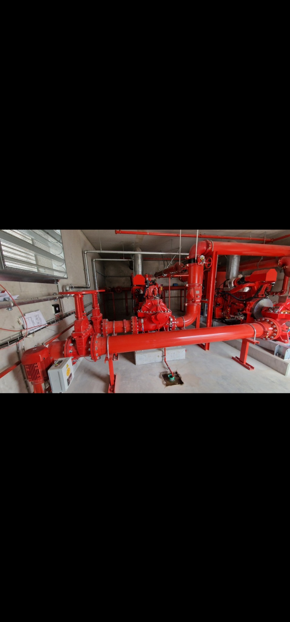 Czerwona instalacja tryskaczowa w pomieszczeniu technicznym. Widoczne rury, zawory, silnik, skrzynka elektryczna i betonowa podstawa. Elementy mocowane do ściany i podłogi.