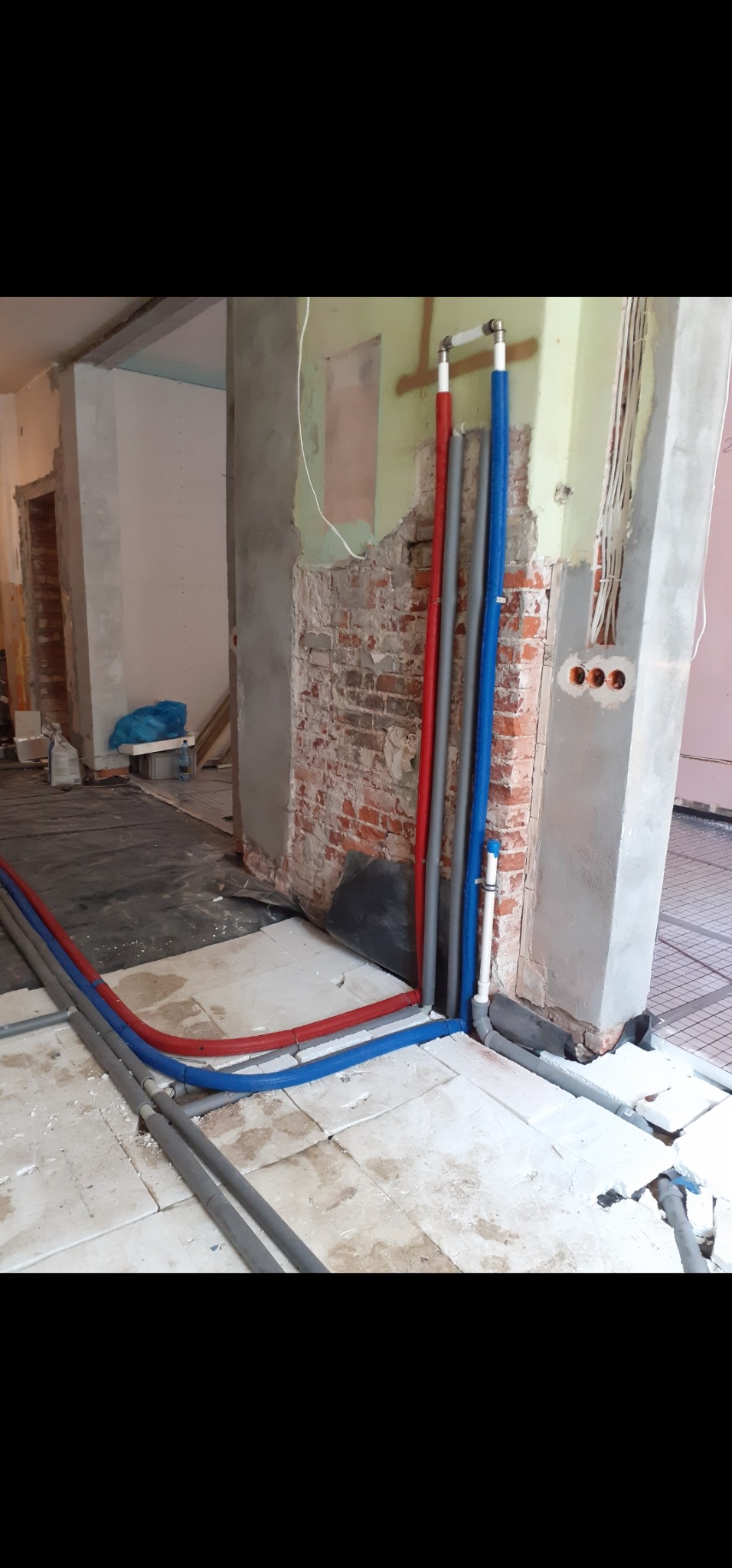 Instalacja rur (czerwona, szara, niebieska) przy ścianie z cegły, na podłodze z płytek. Widoczne przewody elektryczne i elementy konstrukcyjne budynku.