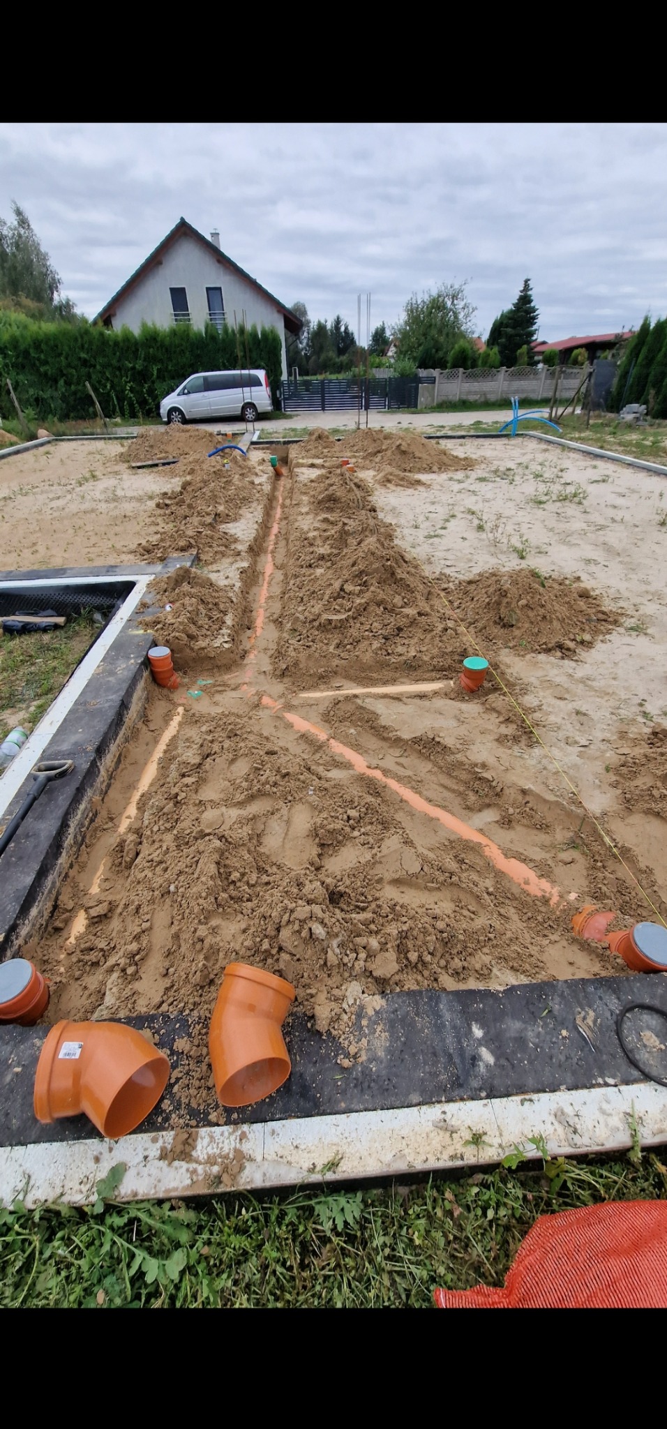 Instalacja kanalizacyjna w gruncie na działce budowlanej, widoczne pomarańczowe rury i studzienki. W tle dom jednorodzinny i zaparkowany van.