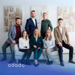 Zespół marketingowy Odado.pl w eleganckich strojach pozuje na kanapie, na tle nowoczesnych obrazów. Uśmiechnięci, w formalnych stylizacjach, gotowi do działania.