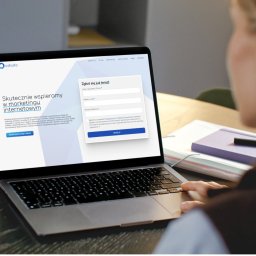 Odado.pl - EKADO GROUP Sp. z o.o. - Laptop z formularzem zgłoszeniowym Odado, marketing internetowy. Widok zza ramienia osoby wpisującej dane. Fioletowy notatnik i długopis obok.