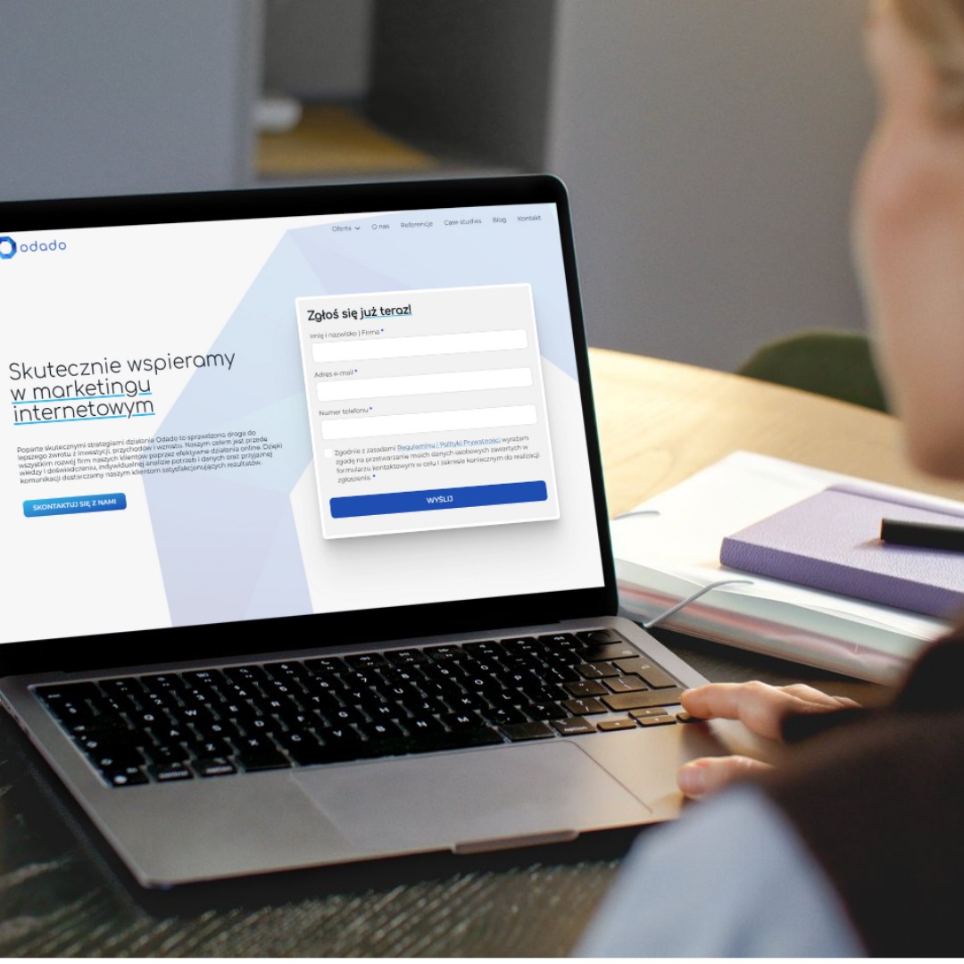 Laptop z formularzem zgłoszeniowym Odado, marketing internetowy. Widok zza ramienia osoby wpisującej dane. Fioletowy notatnik i długopis obok.