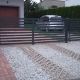 Nowoczesne ogrodzenie panelowe z poziomymi przęsłami w kolorze grafitowym, z bramą wjazdową na posesję wyłożoną kostką i żwirem. W tle dom z garażem.