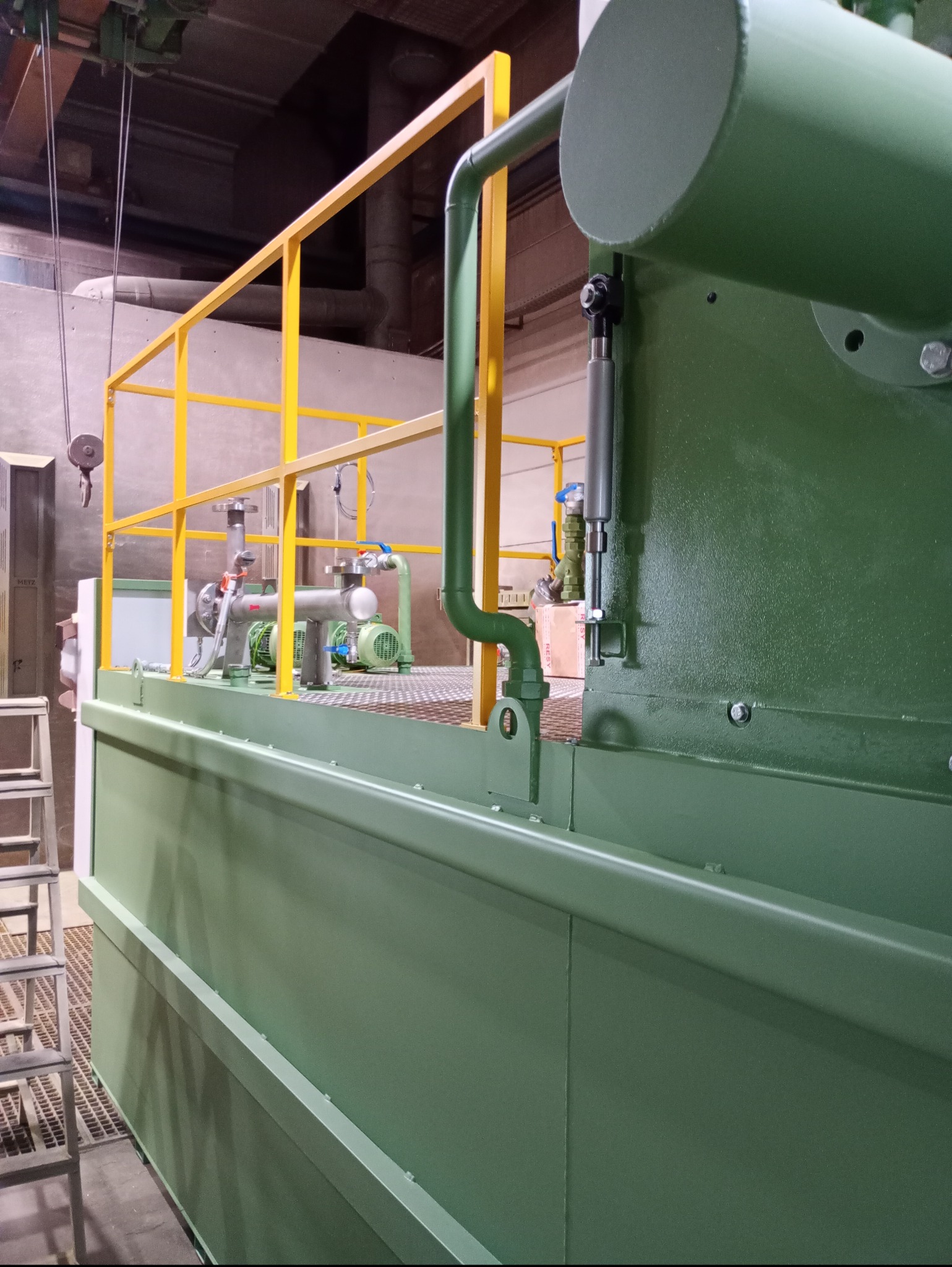 Zielona konstrukcja przemysłowa z żółtymi poręczami, obejmująca pompy i rury. Widoczne elementy złączne i drabina. Ujęcie z dołu, oświetlenie naturalne.