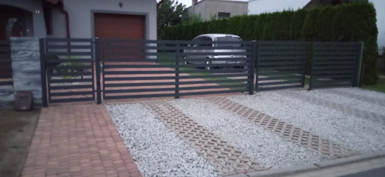 Nowoczesne ogrodzenie panelowe z poziomymi przęsłami w kolorze grafitowym, z bramą wjazdową na posesję wyłożoną kostką i żwirem. W tle dom z garażem.