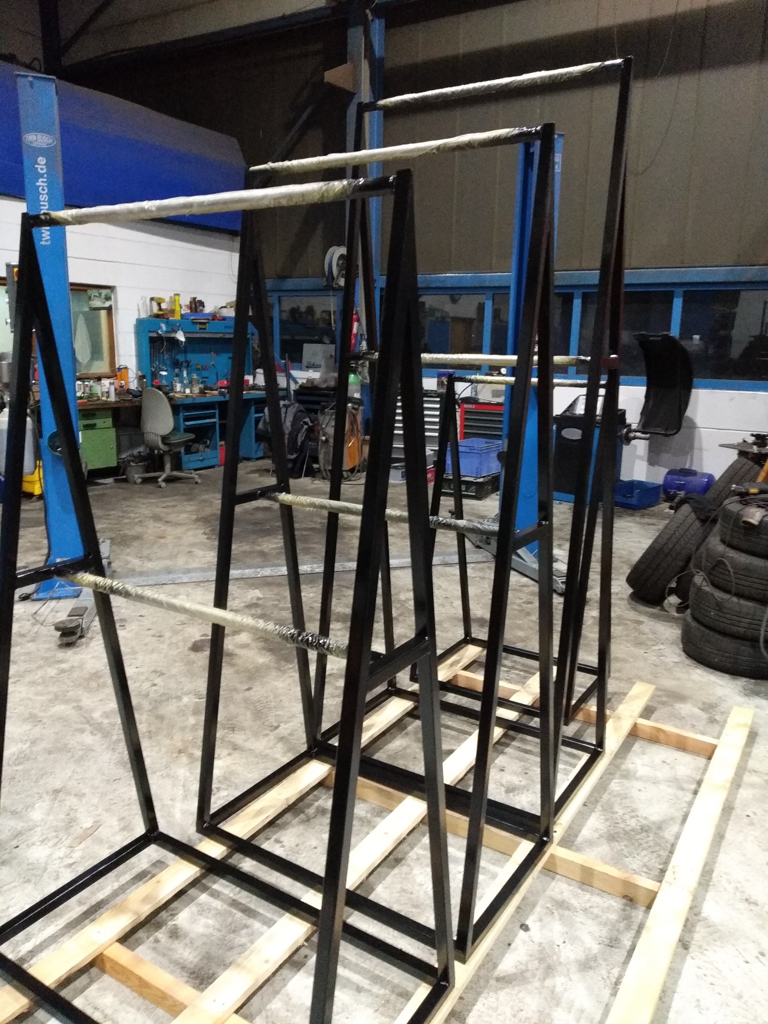 Metalowa konstrukcja schodów z czarnych profili, częściowo owinięta folią, na drewnianej podstawie w warsztacie. Widoczne opony i narzędzia.