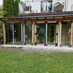 FINESTRA DAWID ZATOR - Zadaszona weranda z drewnianą konstrukcją i przesuwnymi oknami z czarnymi szprosami, widok z zewnątrz na trawnik i zieleń. Częściowo widoczne elementy montażowe.