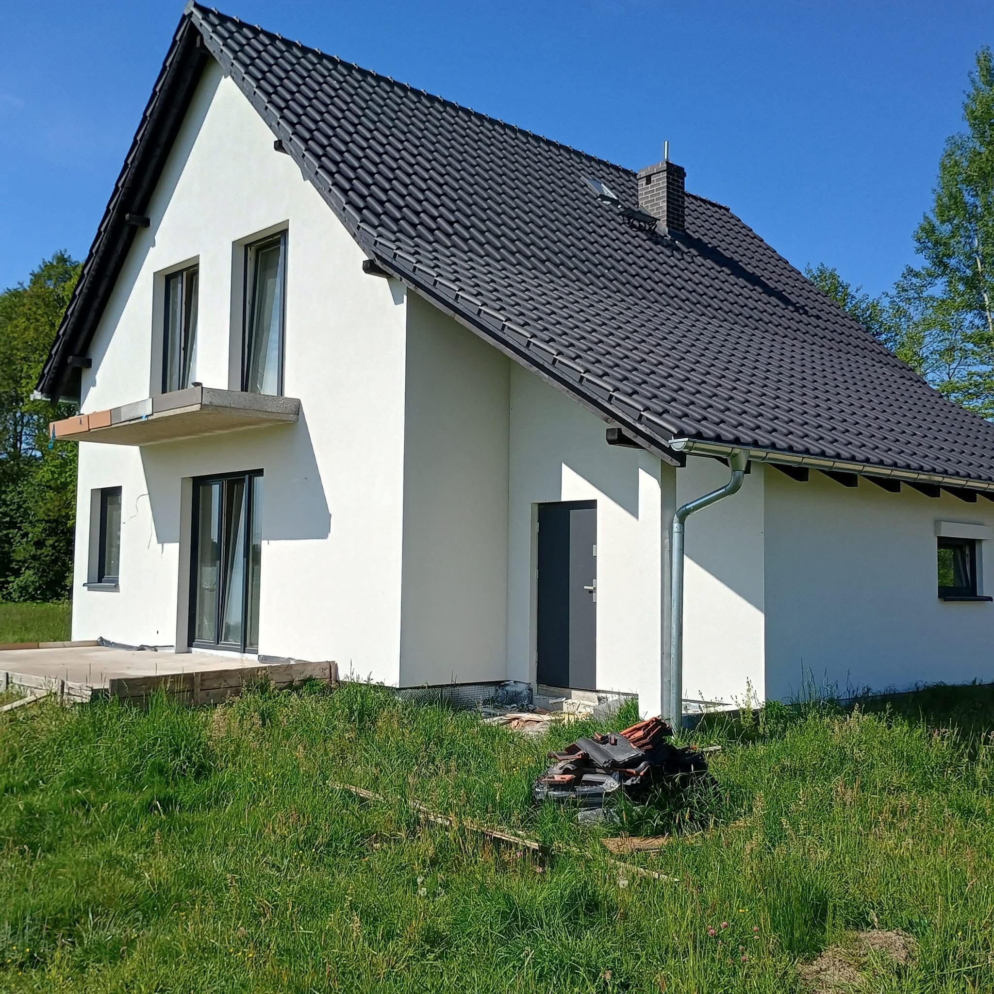Nowoczesny dom jednorodzinny z grafitową dachówką i białymi ścianami, widok z zewnątrz na tle zieleni i błękitnego nieba. Częściowo wykończony balkon i otoczenie.