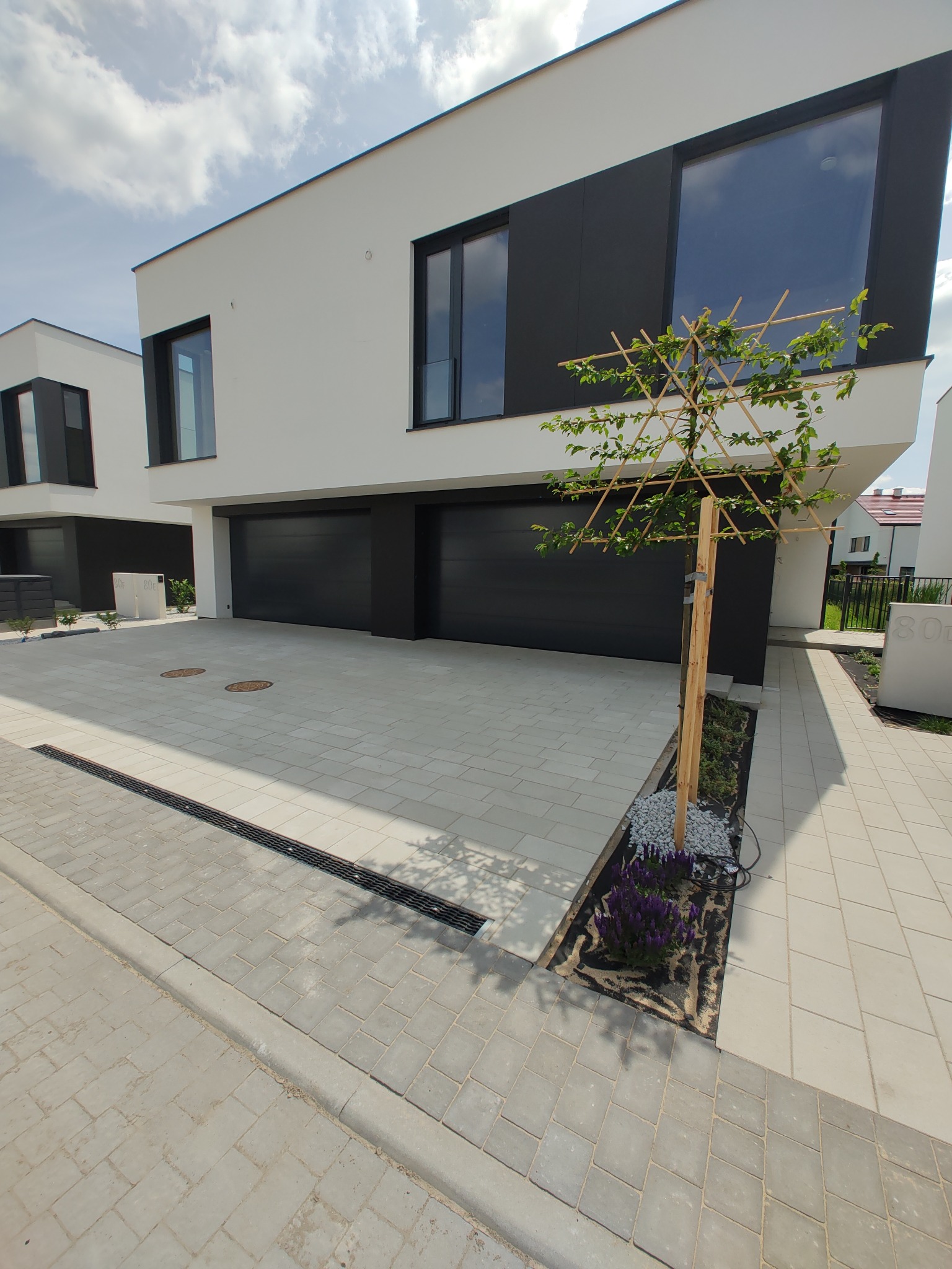 Nowoczesny dom z czarnymi bramami garażowymi i oknami, jasna kostka brukowa na podjeździe, ozdobne drzewko. Minimalistyczny design, słoneczny dzień.