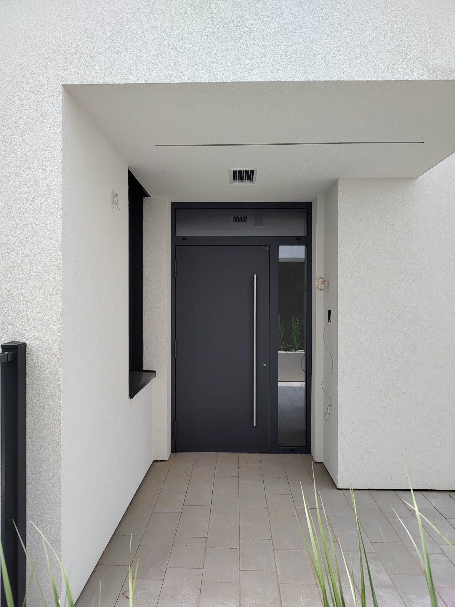 Eleganckie, grafitowe drzwi zewnętrzne z długim, pionowym uchwytem, w nowoczesnym wejściu z szarą podłogą i białymi ścianami. Widoczne okablowanie po prawej.