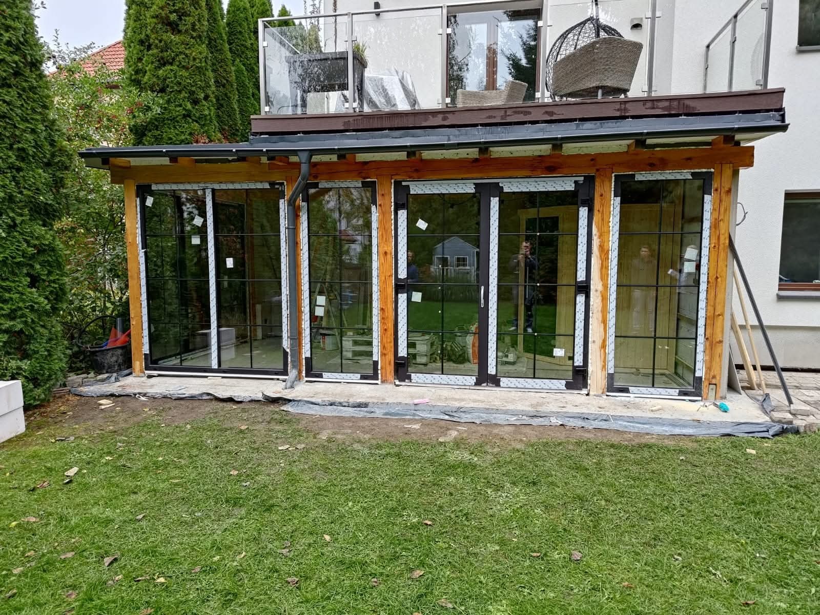 Nowoczesna zabudowa tarasu z drewnianą konstrukcją i dużymi przesuwnymi oknami z czarnymi szprosami. W tle widoczny budynek mieszkalny z balkonem.