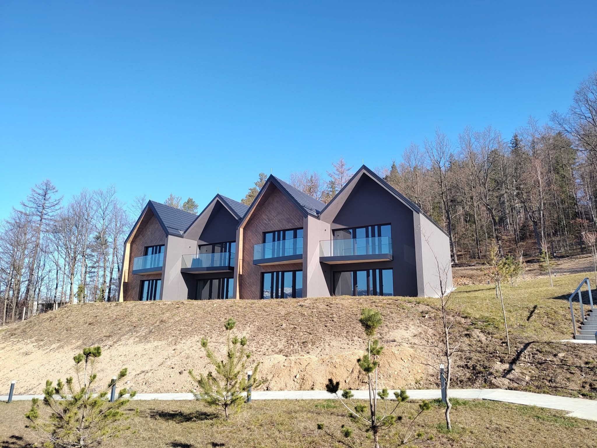 Nowoczesne domy z balkonami z szklanymi balustradami na wzgórzu, widok z dołu, błękitne niebo. Minimalistyczna architektura, drewniane i szare elewacje.