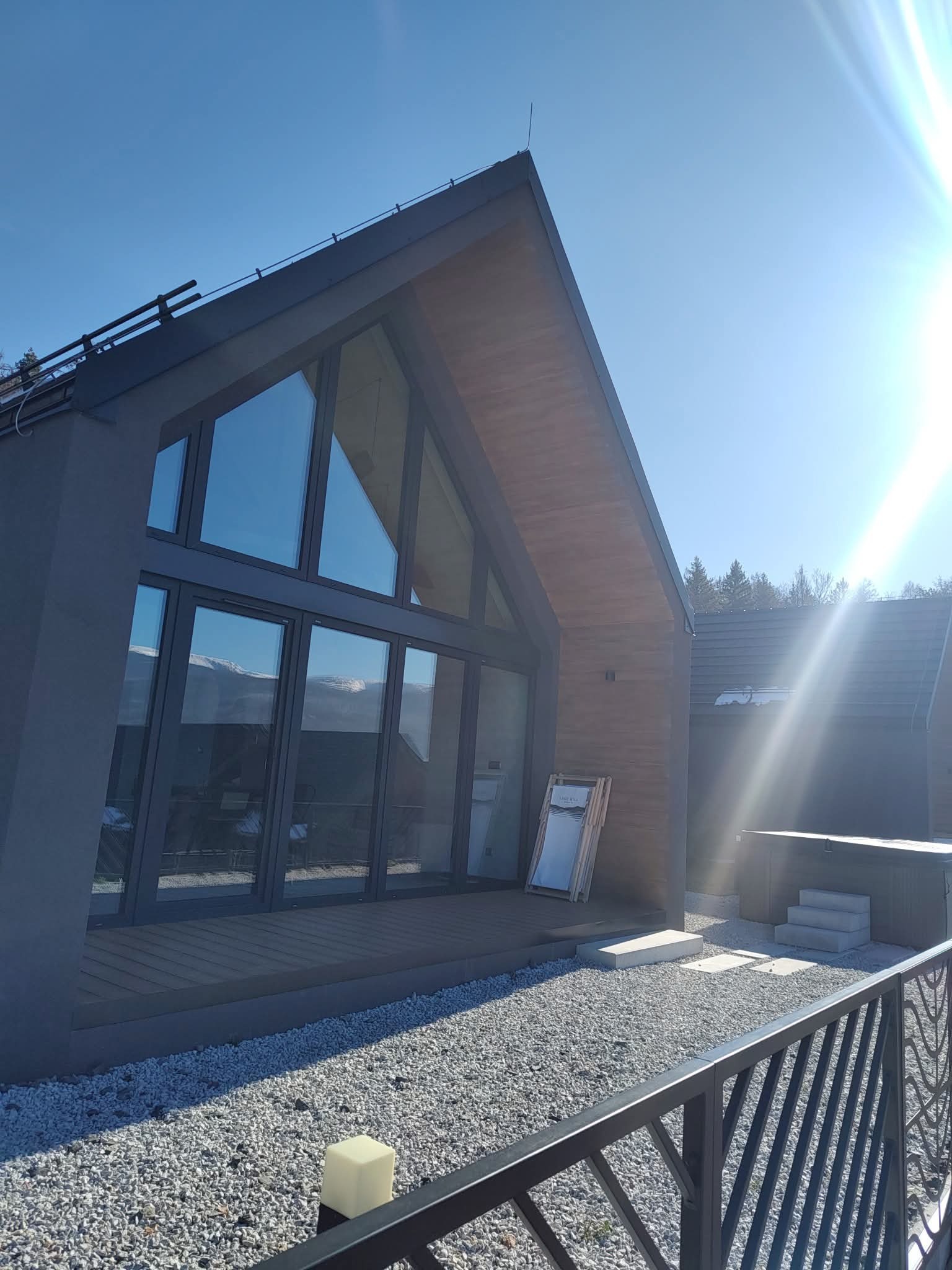 Nowoczesny dom z dużymi oknami aluminiowymi i przesuwnymi drzwiami tarasowymi, widok z zewnątrz na tle błękitnego nieba i gór. Minimalistyczna architektura.