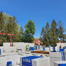 Wznoszenie ścian domu z bloczków silikatowych, widoczni pracownicy w kaskach i kamizelkach odblaskowych, czerwony dźwig budowlany w tle, palety z materiałem budowlanym z widocznym logo producenta.