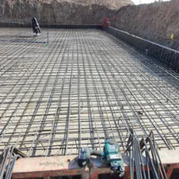 Siatka zbrojeniowa na przygotowanym podłożu, widoczne narzędzia zbrojarskie i szalunki, grunt w tle.