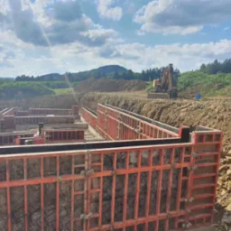 Przygotowanie szalunków pod wylewkę fundamentów na budowie, widoczny wykop i częściowo zabetonowana podstawa, w tle koparka i krajobraz zalesionych wzgórz pod błękitnym, obłocznym niebem.