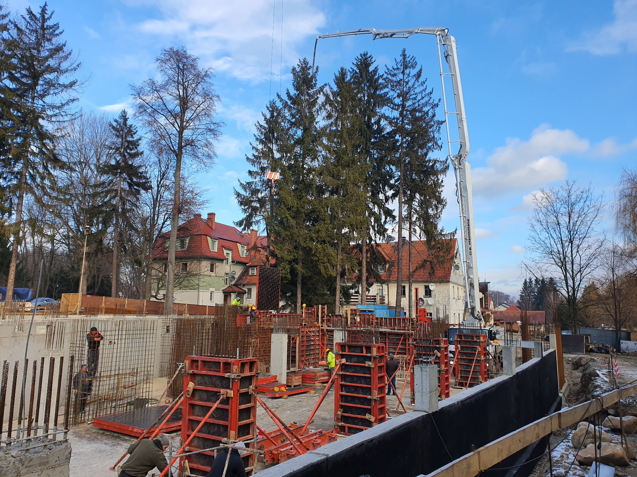 Wykop pod budynek z częściowo zabetonowanymi ścianami, szalunkami i zbrojeniami; w tle budynki mieszkalne i wysięgnik pompy do betonu.