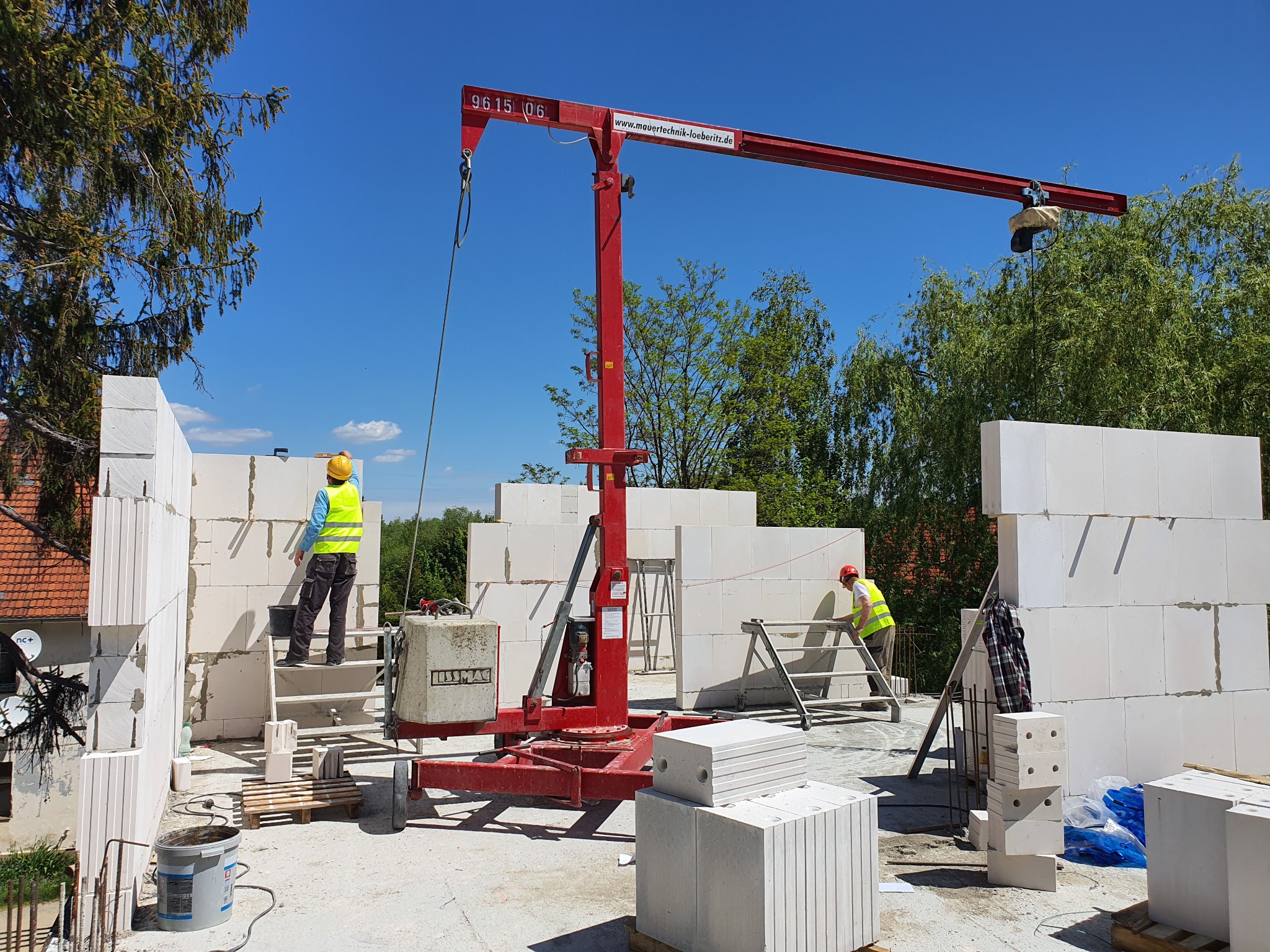 Budowa domu z białych bloczków, widoczny dźwig budowlany, pracownicy w kaskach i kamizelkach odblaskowych, palety z bloczkami, etap wznoszenia ścian.