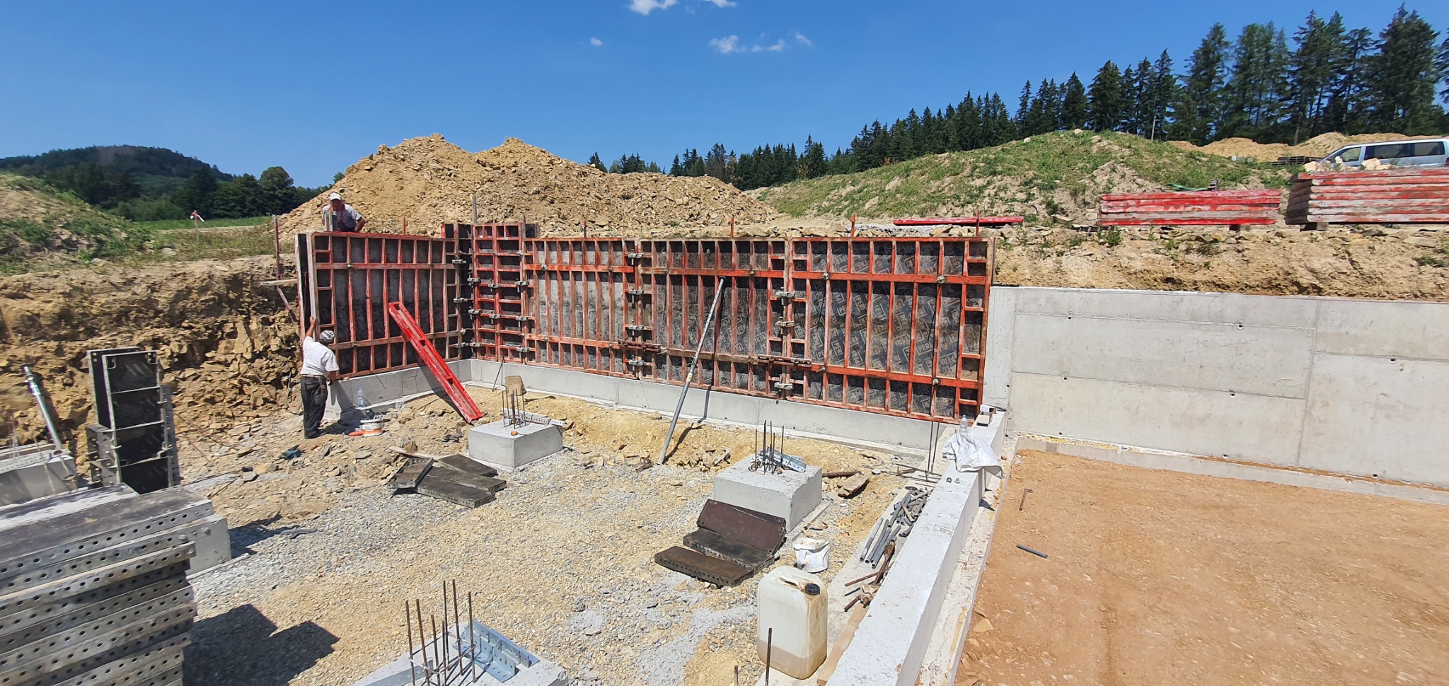Budowa ściany oporowej: szalunek z czerwonymi ramami, betonowe fundamenty, pracownicy na placu budowy, w tle wzgórze z drzewami.