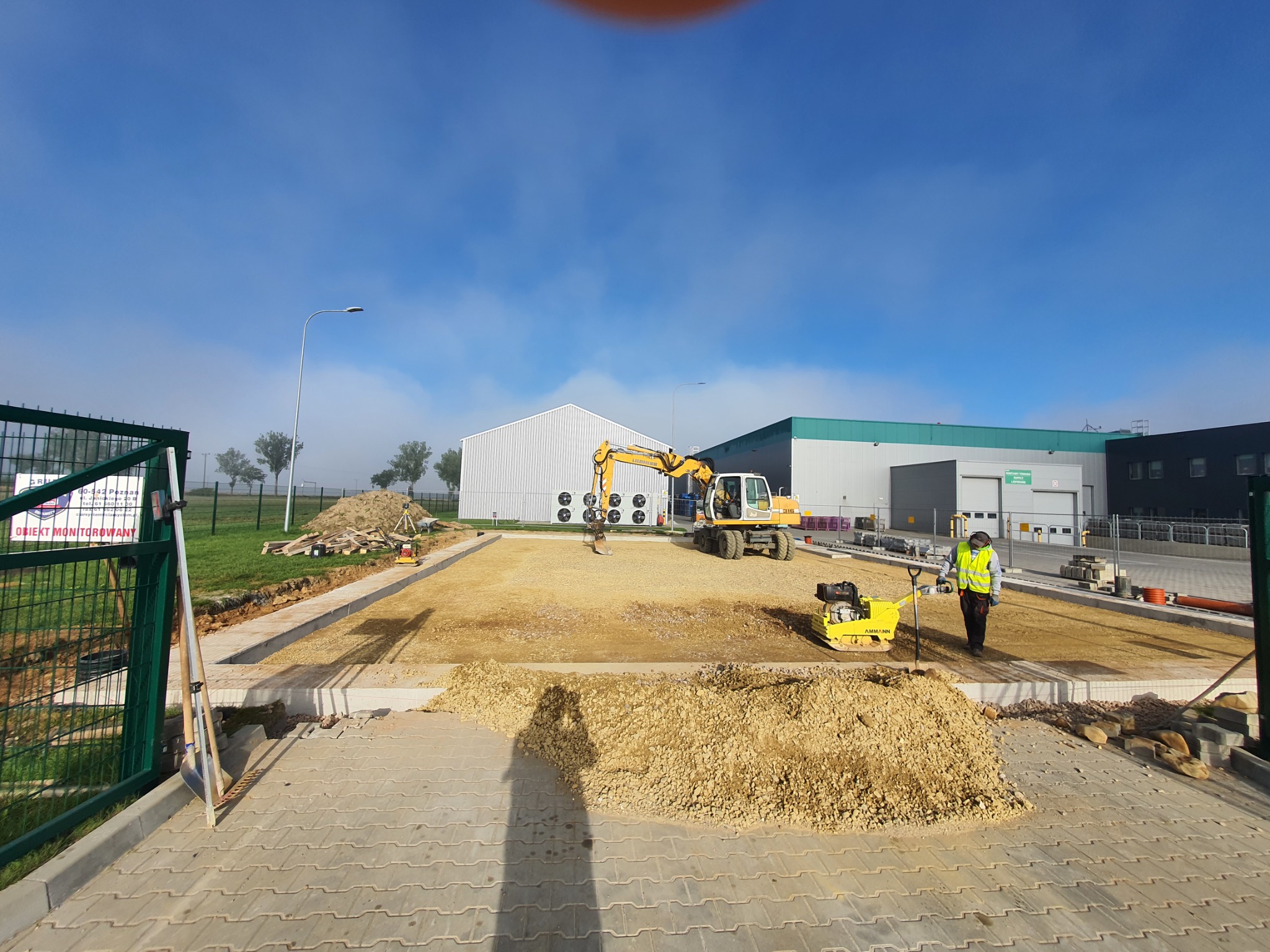 Przygotowanie terenu pod budowę hali: żółta koparka Liebherr 316, ubijarka Ammann i pracownik w kamizelce odblaskowej na placu budowy, widok zza ogrodzenia.